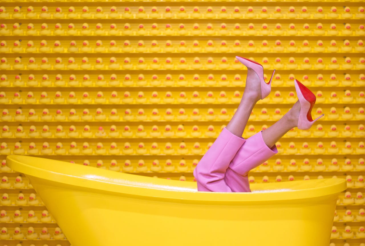 about-02 Legs in pink pants and red high heels pop out of a yellow bathtub against a creative rubber duck wall.